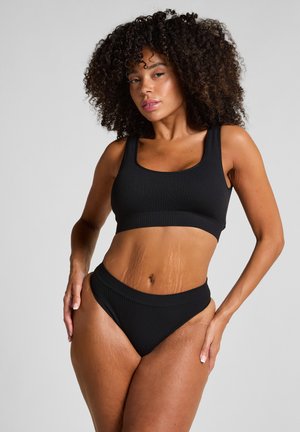 Donna con capelli ricci che indossa un crop top a costine nero e biancheria intima a vita alta, in piedi con una mano sul fianco su uno sfondo neutro.
