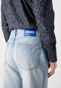 Jeans in denim azzurro con vestibilità dritta, dotati di un'etichetta blu con la scritta "HUGO". Il tessuto ha una consistenza morbida e lievi effetti di scolorimento.