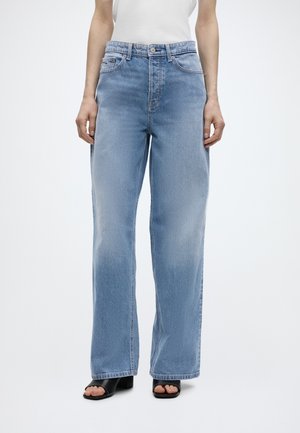 Osoba w jasnoniebieskich jeansach z szerokimi nogawkami, białym topie bez rękawów i czarnych sandałach na słupku z odkrytymi palcami na białym tle.