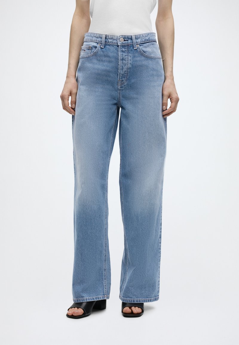 Άτομο που φοράει ανοιχτό μπλε τζιν παντελόνι με φαρδιά μπατζάκια, λευκό αμάνικο τοπ και μαύρα πέδιλα με ανοιχτά δάχτυλα και χοντρό τακούνι σε λευκό φόντο.