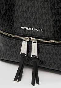 Zaino nero con motivo, caratterizzato da una finitura lucida, accessori in argento, doppie cerniere con nappine e il logo "Michael Kors" in evidenza.