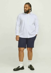 White button-up shirt, navy shorts, black loafers, and white socks with decorative lettering. Model is standing against a light background.