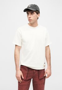 Νεαρός άνδρας που φοράει εκρού κοντομάνικο T-shirt, μπορντό παντελόνι τύπου cargo και σκουρόχρωμο γκρι καπέλο μπέιζμπολ, κοιτάζει προς τα πλάγια μπροστά σε ουδέτερο φόντο.