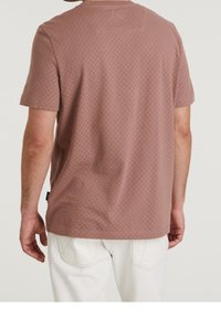 Mężczyzna w brązowej, krótkiej koszuli o fakturze i białych spodniach, widoczny od tyłu na tle jednolitym.