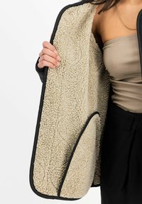 Veste noire à couche extérieure avec doublure en polaire beige douce. Présente un col rond et un design minimaliste, tenue ouverte pour dévoiler la texture intérieure.