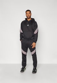 Sort hættetrøje og bukser i sæt med hvide og røde diagonale striber på ærmerne og benene, samt et Adidas-logo på brystet.