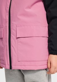 Veste rose et noire dotée d'une fermeture éclair à l'avant, de deux grandes poches à rabat et d'une texture lisse, associée à un jogging gris.