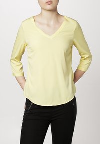 Blouse jaune à col en V avec des manches trois-quarts, texture lisse et ourlet arrondi. Associée à un pantalon noir avec des accents zippés.
