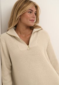 Beige stickad tröja med halvdragkedja, med ett texturerat mönster och en guldfärgad dragkedja, designad för avslappnad användning.