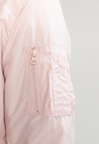 Světle růžová bomber bunda s texturovanou tkaninou, která má zipovou kapsu na rukávu a našité detaily pro zvýraznění.