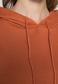 Terakotovou mikinu s měkkou strukturou, která má kapuci se stahovací šňůrkou a žebrované detaily u výstřihu. Jednoduchý, neformální design.