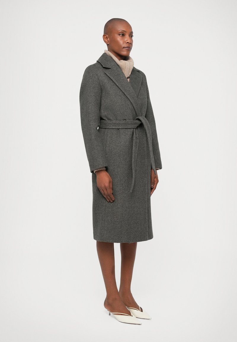 Grauer Wollumhang mit einem Gürtel, Reverskragen, langen Ärmeln und Seitentaschen. Tragen zu einem beigen Rollkragenpullover und weißen Absätzen.