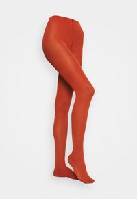 Jambes portant des collants opaques couleur orange brûlé, posées avec un pied à plat et l'autre légèrement relevé sur un fond clair.