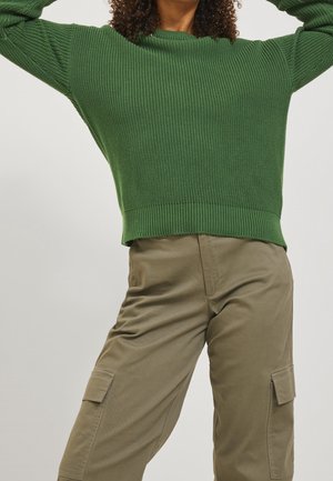 Personne portant un pull en maille vert et un pantalon cargo beige, posant les bras levés, sur un fond clair uni.