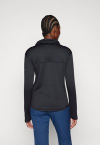 Camisa negra de manga larga con un acabado texturizado, cuello alto y dobladillo ligeramente curvado. Presenta puños extendidos con agujeros para los pulgares.