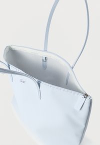 Jasnoniebieska torba typu tote marki Lacoste z podwójnymi uchwytami, zamkiem błyskawicznym na górze i haftowanym logo krokodyla, pokazana otwarta, odsłaniająca wewnętrzną lamówkę z logo marki.
