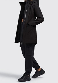 Sort vandtæt jakke med hætte, med lynlås foran, to sidelommer og et slankt design, parret med sorte cargobukser og sneakers.