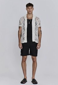 Bijela čipkasta košulja s kratkim rukavima preko crnog topa, uparen s crnim cargo šortsevima i crnim natikačama. Model stoji ispred neutralne pozadine.