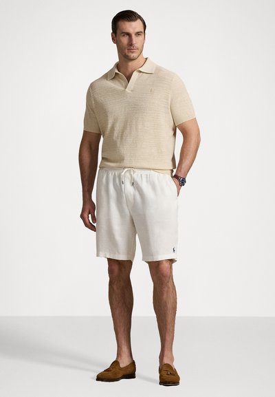 Beige pletené polo tričko s krátkým rukávem, kombinováno s bílými lněnými šortkami s elastickým pasem a malým vyšitým logem. Hnědé mokasíny.
