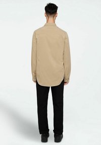 Beige long-sleeve shirt with a curved hem, buttoned cuffs, and back yoke, worn with black pants and black shoes.