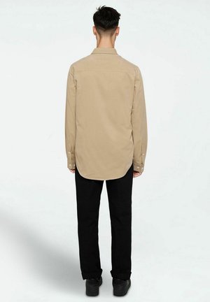 Beige long-sleeve shirt with a curved hem, buttoned cuffs, and back yoke, worn with black pants and black shoes.