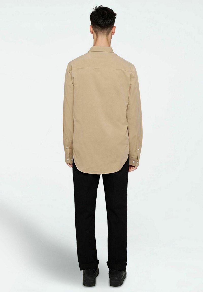 Beige long-sleeve shirt with a curved hem, buttoned cuffs, and back yoke, worn with black pants and black shoes.