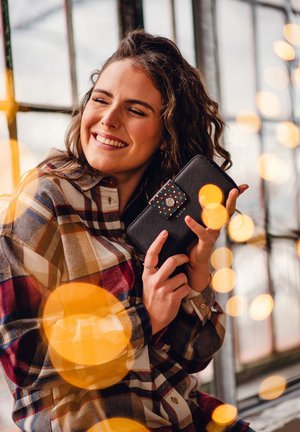 Black wallet with a patterned flap featuring colorful dots. The material appears textured. The background includes blurred light spots.