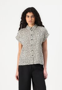 Camicia a maniche corte color crema con un motivo astratto nero, con colletto a bottoni e un taglio morbido. Il materiale è liscio e leggero.