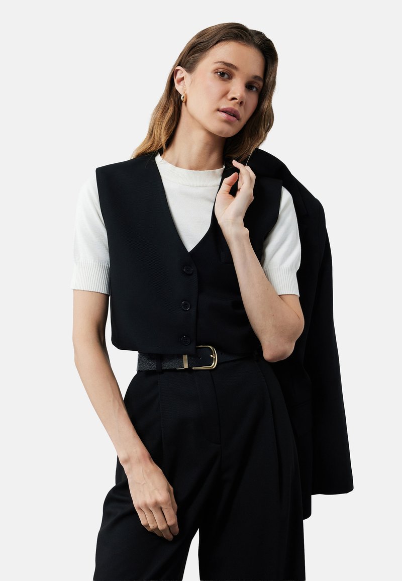 Zwarte vest over een wit shirt met korte mouwen, voorzien van een v-hals en knopen. Gecombineerd met hoge zwarte broek en een zwarte riem met een gouden gesp.