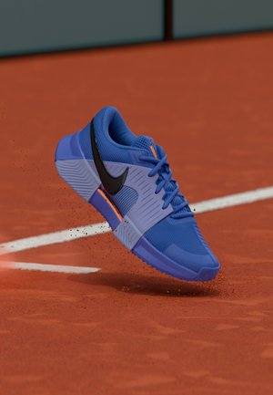 Chaussure de sport bleue avec dessus en mesh, logo swoosh noir, semelle texturée et accents orange, montrée en l'air au-dessus d'une surface de terrain rouge.