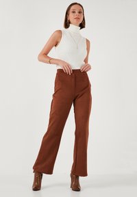 Braune Wildleder-Hosen im weiten Schnitt, kombiniert mit einem weißen gerippten ärmellosen Rollkragenpullover. Braune Stiefeletten sind am unteren Ende sichtbar. Einfaches Design, figurschmeichelnde Passform.