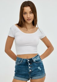 Camiseta blanca recortada con escote ancho, combinada con pantalones cortos de mezclilla azul de tiro alto que presentan un cierre de botón y un ligero desgastado.