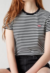 Personne aux longs cheveux roux portant un t-shirt Levi's rayé noir et blanc et un short noir, assise les bras croisés devant.