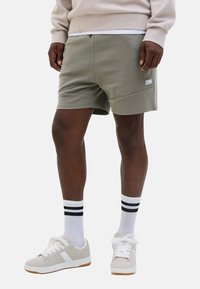 Man in een lichtgrijze short, witte sokken met zwarte strepen, beige sneakers en een beige sweatshirt, staand tegen een witte achtergrond.
