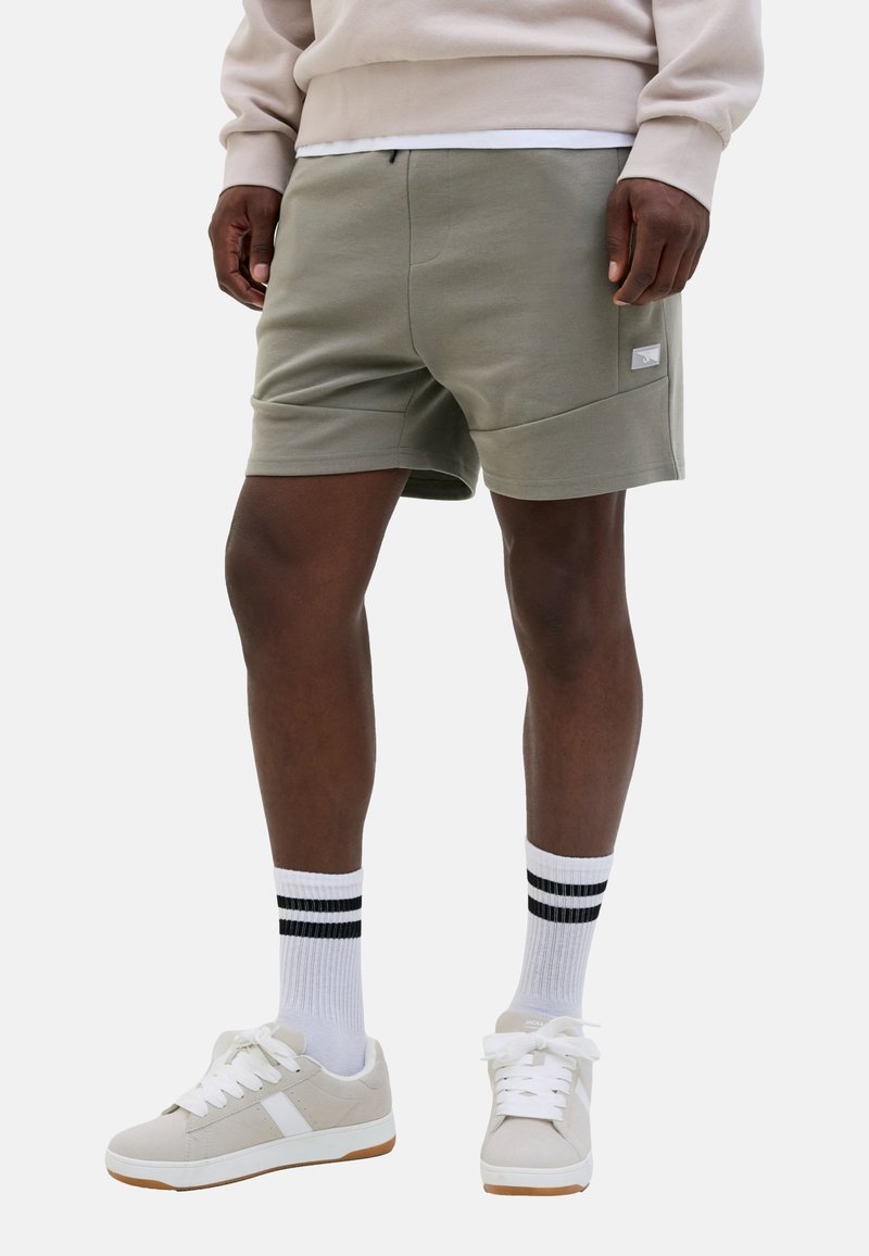 Man in een lichtgrijze short, witte sokken met zwarte strepen, beige sneakers en een beige sweatshirt, staand tegen een witte achtergrond.
