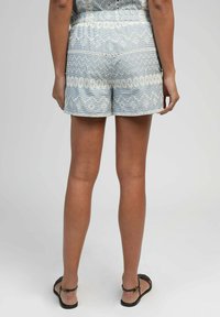 Femme portant un short à motifs bleu clair et des sandales à bride de cheville noires, se tenant contre un fond gris uni.