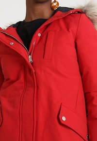 Rote Parkajacke mit einem mit Fell gefütterten Kapuze, ausgestattet mit einem Reißverschluss und Druckknopfverschluss, seitlichen Taschen und einer glatten Textur.