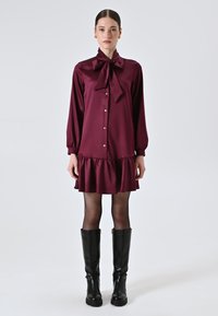 Bordeauxfarbenes Satinhemdkleid mit einer Schleifenkragen, Knopfleiste, Ballonärmeln und einem Rüschensaum. Kombiniert mit kniehohen schwarzen Stiefeln.