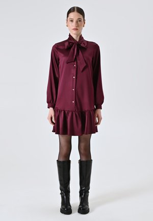 Bordeauxfarbenes Satinhemdkleid mit einer Schleifenkragen, Knopfleiste, Ballonärmeln und einem Rüschensaum. Kombiniert mit kniehohen schwarzen Stiefeln.