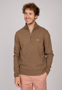 Sweater zippé marron en maille texturée, doté d'un col montant et d'un ourlet côtelé, porté sur une chemise beige clair.