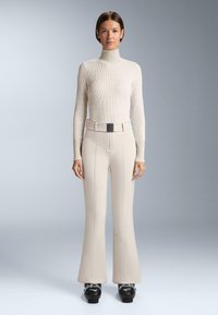 Beżowy prążkowany sweter z golfem zestawiony z wysokim stanem dzwonowatych spodni z pionowymi przeszyciami i czarnym paskiem z klamrą. Czarne buty na platformie.