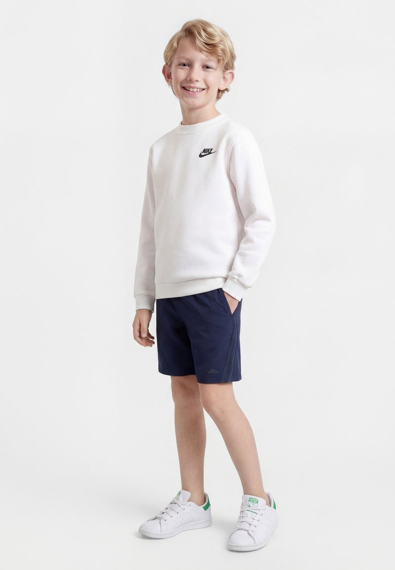 Garçon souriant aux cheveux blonds portant un sweat-shirt blanc Nike, un short bleu marine et des baskets blanches avec des accents verts, debout devant un fond uni.