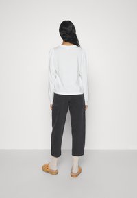 Camisa blanca de manga larga con hombros abullonados, combinada con pantalones negros recortados. Zuecos marrones y calcetines beiges completan el atuendo. Diseño sencillo.