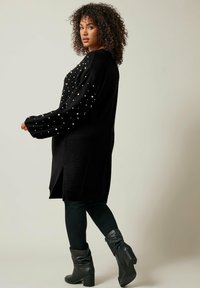 Maglione nero lavorato a maglia con decorazioni in perle sulle maniche, caratterizzato da una vestibilità morbida, fessure laterali, indossato sopra pantaloni neri aderenti e stivali al ginocchio.