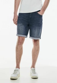 Muž oblečený v světlých denimových kraťasech, bílých teniskách a bílém tričku, stojící s jednou rukou v kapse na bílém pozadí.