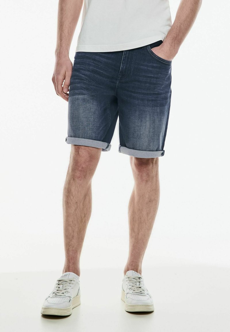 Muž oblečený v světlých denimových kraťasech, bílých teniskách a bílém tričku, stojící s jednou rukou v kapse na bílém pozadí.