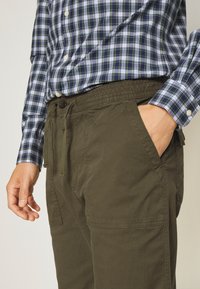 Pantalones verde oliva con cintura elástica y cordón, con bolsillos laterales y un corte relajado, combinados con una camisa de cuadros azul.