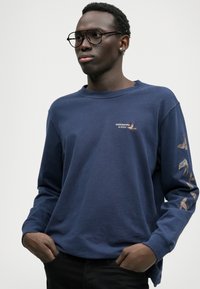 Tume sinine pika varrukaga T-särk, mis on valmistatud puuvillast, millel on rinnal väike logo ja värviline linnu tikand varrukatel.