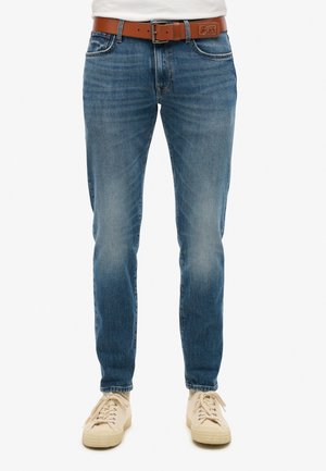 Vaqueros de mezclilla azul con corte slim, lavado desteñido y un cinturón de cuero marrón. Presentan un diseño clásico de cinco bolsillos y detalles de costura sutiles.
