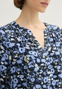 Niebieska bluzka w kwiatowy wzór z dekoltem w kształcie litery V, z przodem na guziki i lekkim materiałem w kolorach akcentujących białym i żółtym.